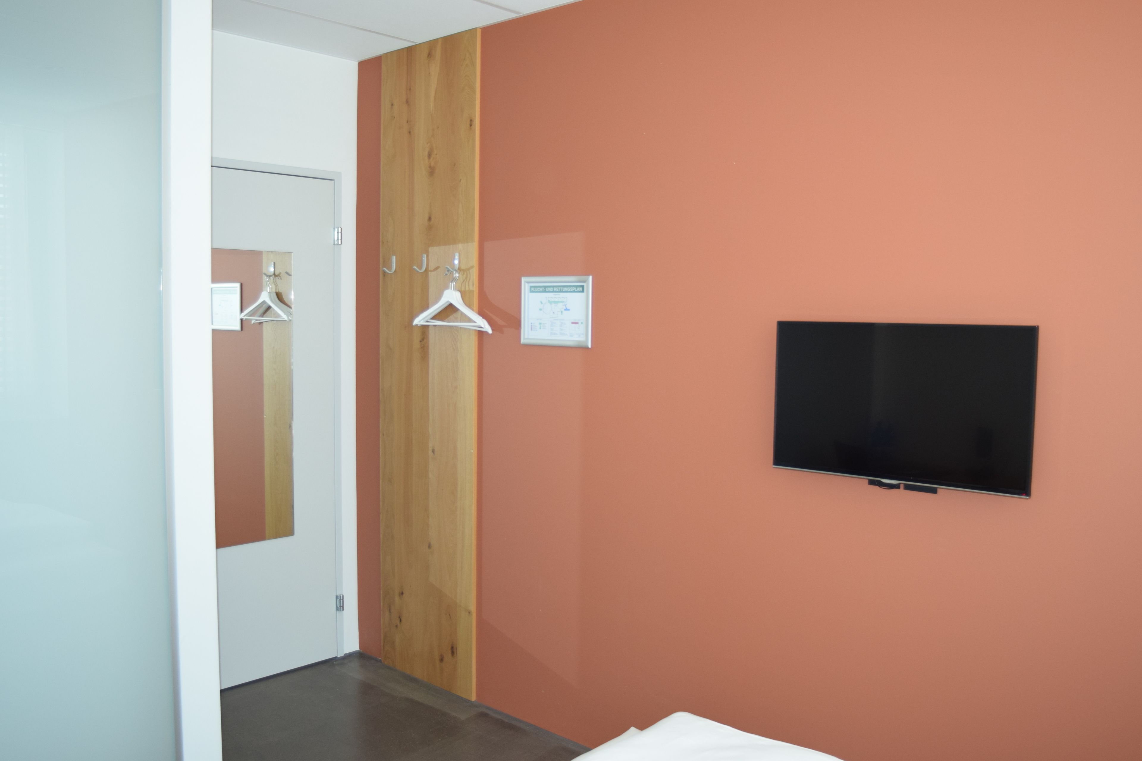 A hotel room with a television mounted on the wall, a wooden panel on one wall, and a door with a mirror and clothes hanger.