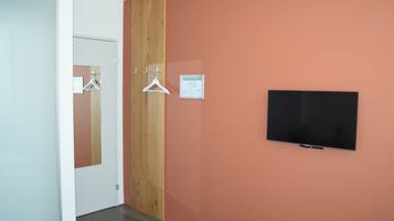 A hotel room with a television mounted on the wall, a wooden panel on one wall, and a door with a mirror and clothes hanger.