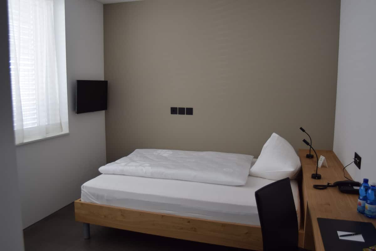 Un lit simple avec un cadre en bois, un bureau avec une chaise, un téléviseur à écran plat fixé au mur et une fenêtre avec des stores.