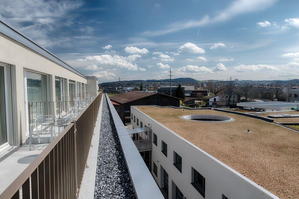 Un bâtiment moderne doté d’un long balcon, d’une fontaine circulaire et offrant une vue sur un paysage urbain en arrière-plan.