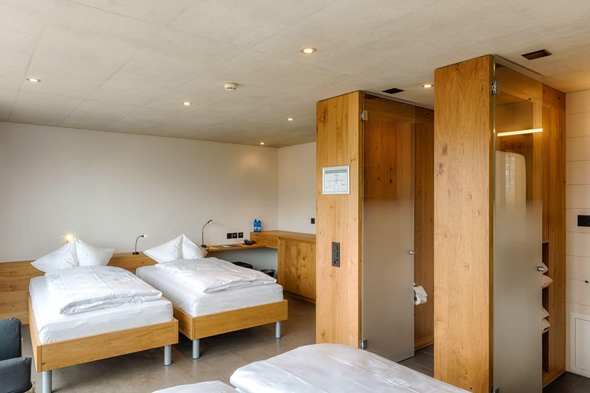 Une chambre d’hôtel avec deux lits simples, un bureau en bois, une chaise, une armoire et une petite kitchenette.