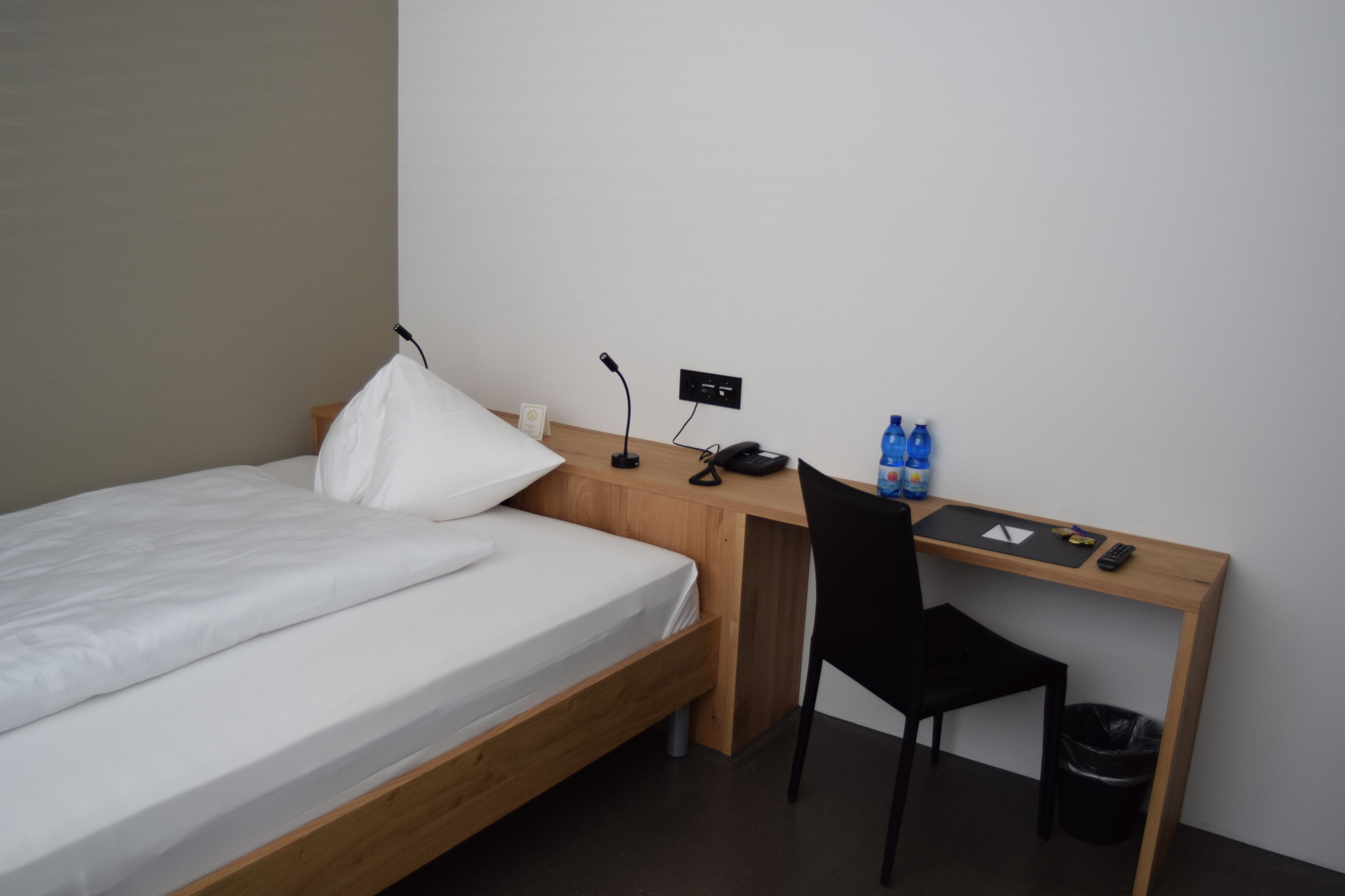 A single bed with a wooden frame, a desk with a chair, a phone, a trash bin, and bottled water.