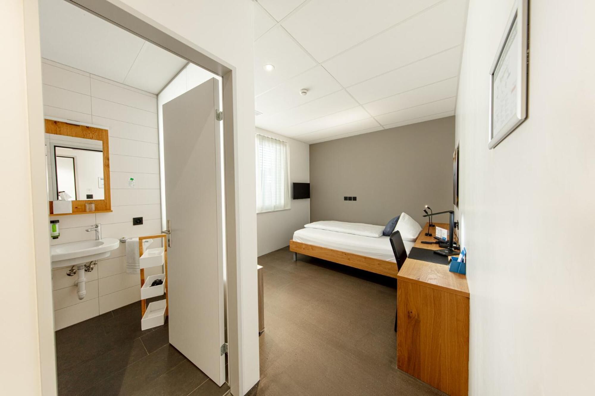 A modern hotel room with a bathroom, a bed, a desk, and a TV.