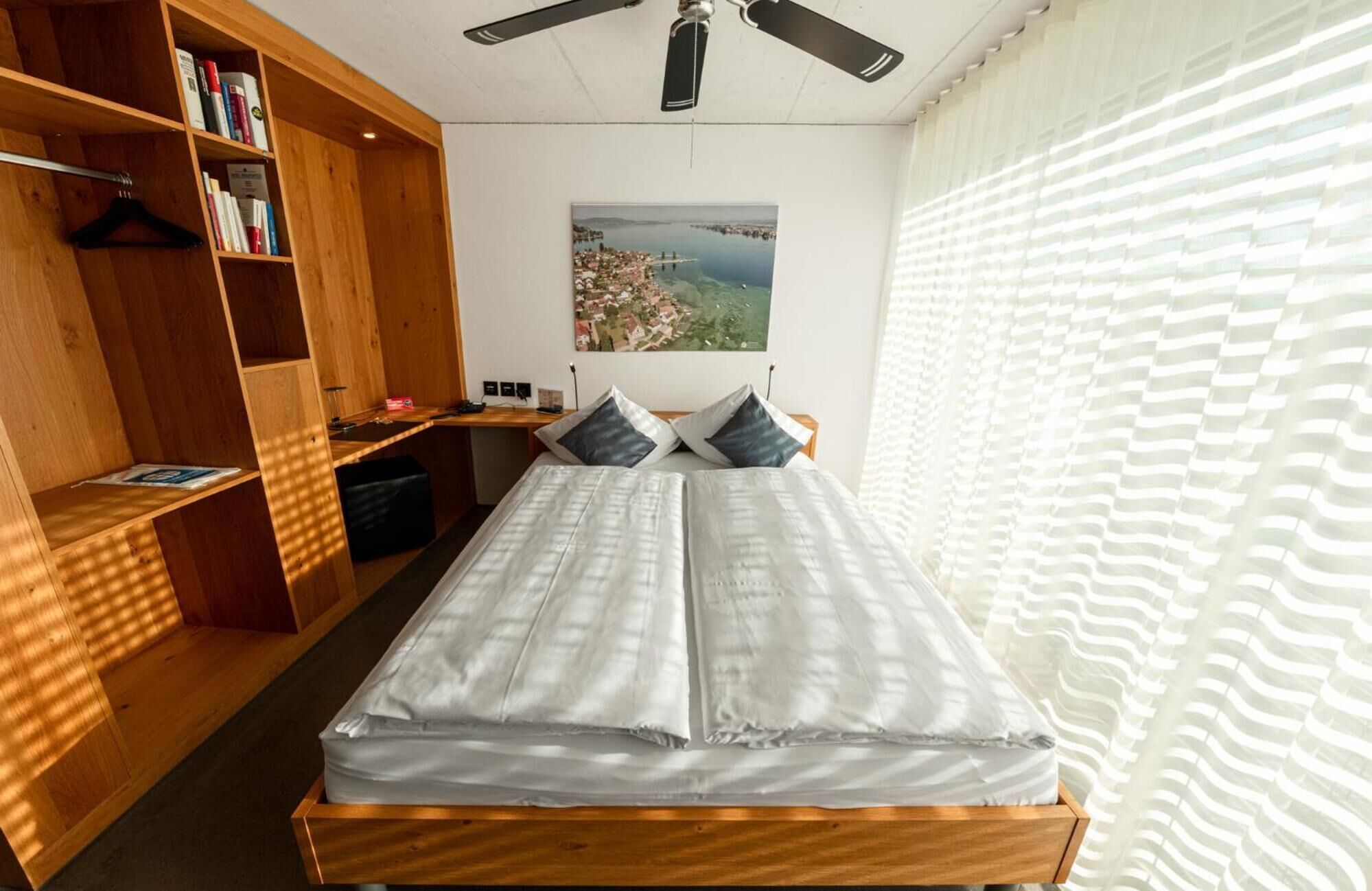 Ein Schlafzimmer mit einem Bett, einem Holzregal, einem Schreibtisch, einem Deckenventilator und einem Fenster mit Jalousien.