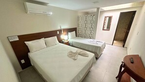 Quarto de hotel com uma cama grande, uma cama menor, uma mesinha de cabeceira de madeira com abajur, um biombô decorativo e uma mesa de madeira com controle remoto.