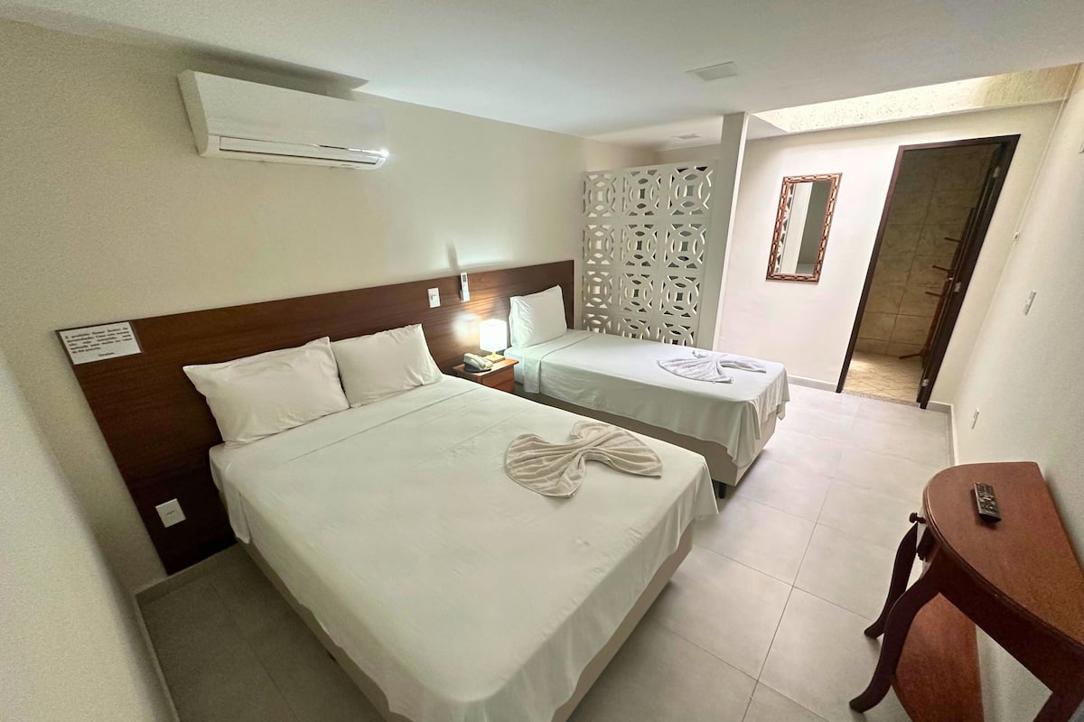 A hotel room with a large bed, a smaller bed, a wooden nightstand, a desk, and an air conditioning unit.