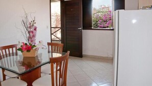 Uma cozinha compacta com geladeira branca, uma mesa de jantar de madeira com cadeiras, e uma janela com flores cor-de-rosa.
