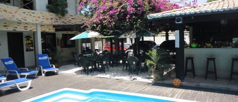 A pool area with lounge chairs, a bar, and a covered seating area with a flowering tree.