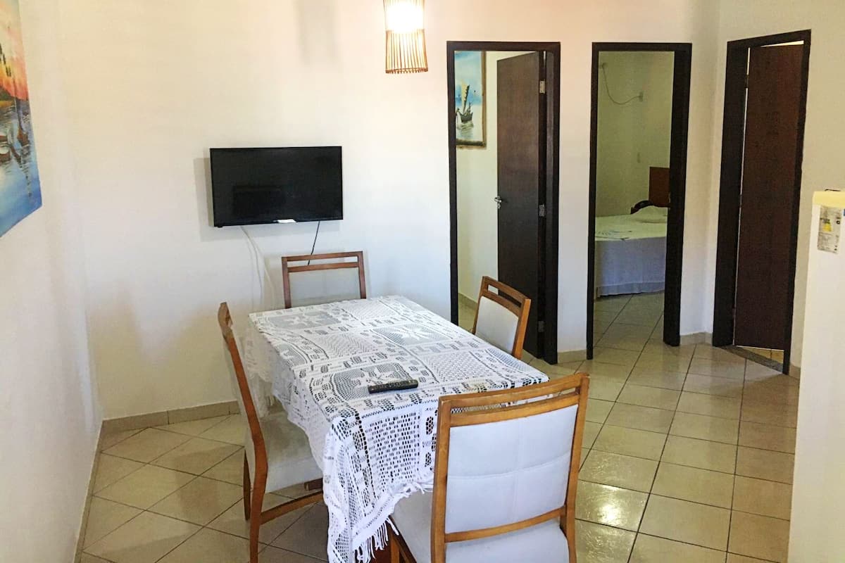 A dining area with a table covered by a lace tablecloth, wooden chairs, a television mounted on the wall, and a door leading to another room.