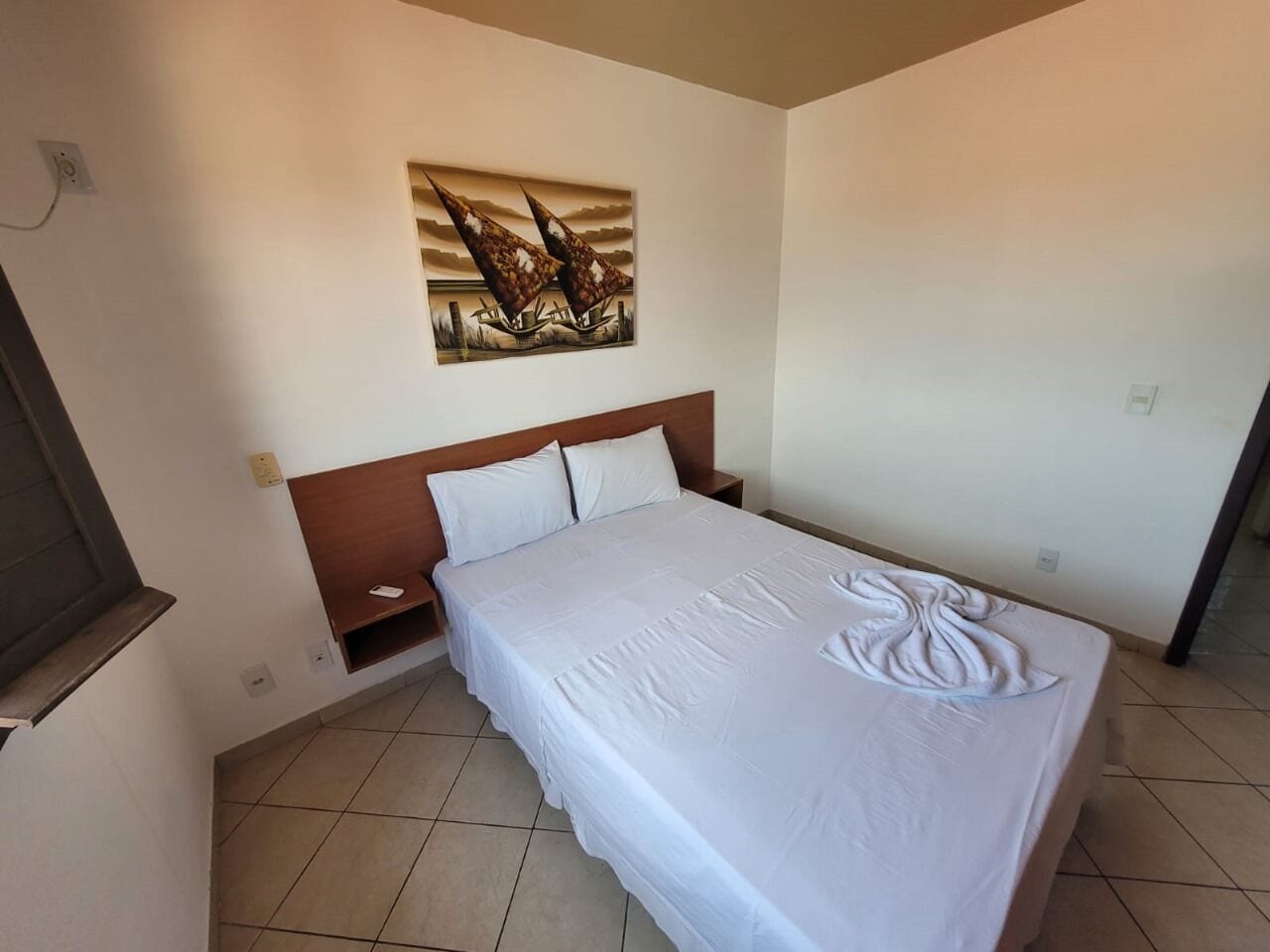 A neatly made bed with a white bedspread and pillows, a wooden headboard, a bedside table, and a wall-mounted painting.
