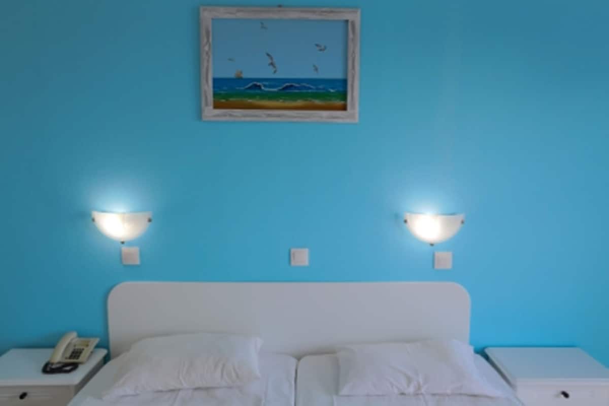 A room with a bed, two bedside tables, a painting of a beach scene, and wall-mounted lights.