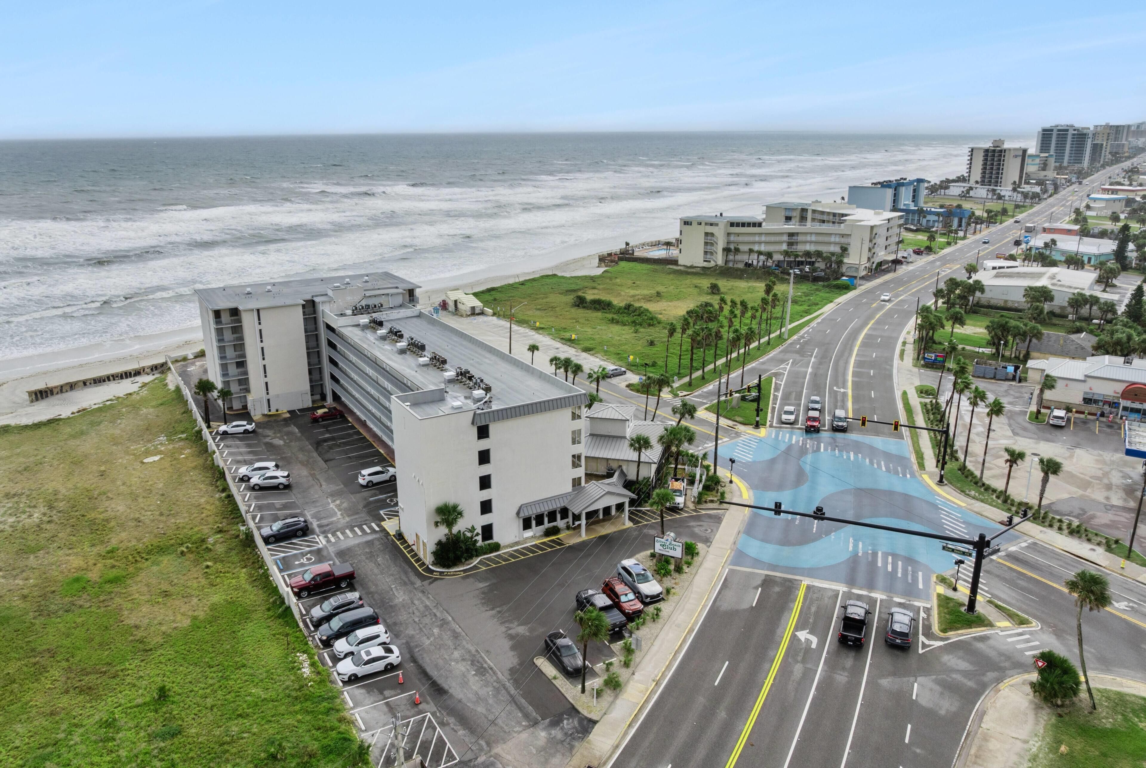An aerial view of a coastal hotel with a parking lot, a road with traffic lights, and a beach in the background.