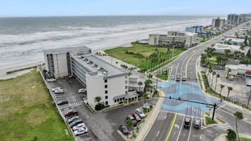 An aerial view of a coastal hotel with a parking lot, a road with traffic lights, and a beach in the background.
