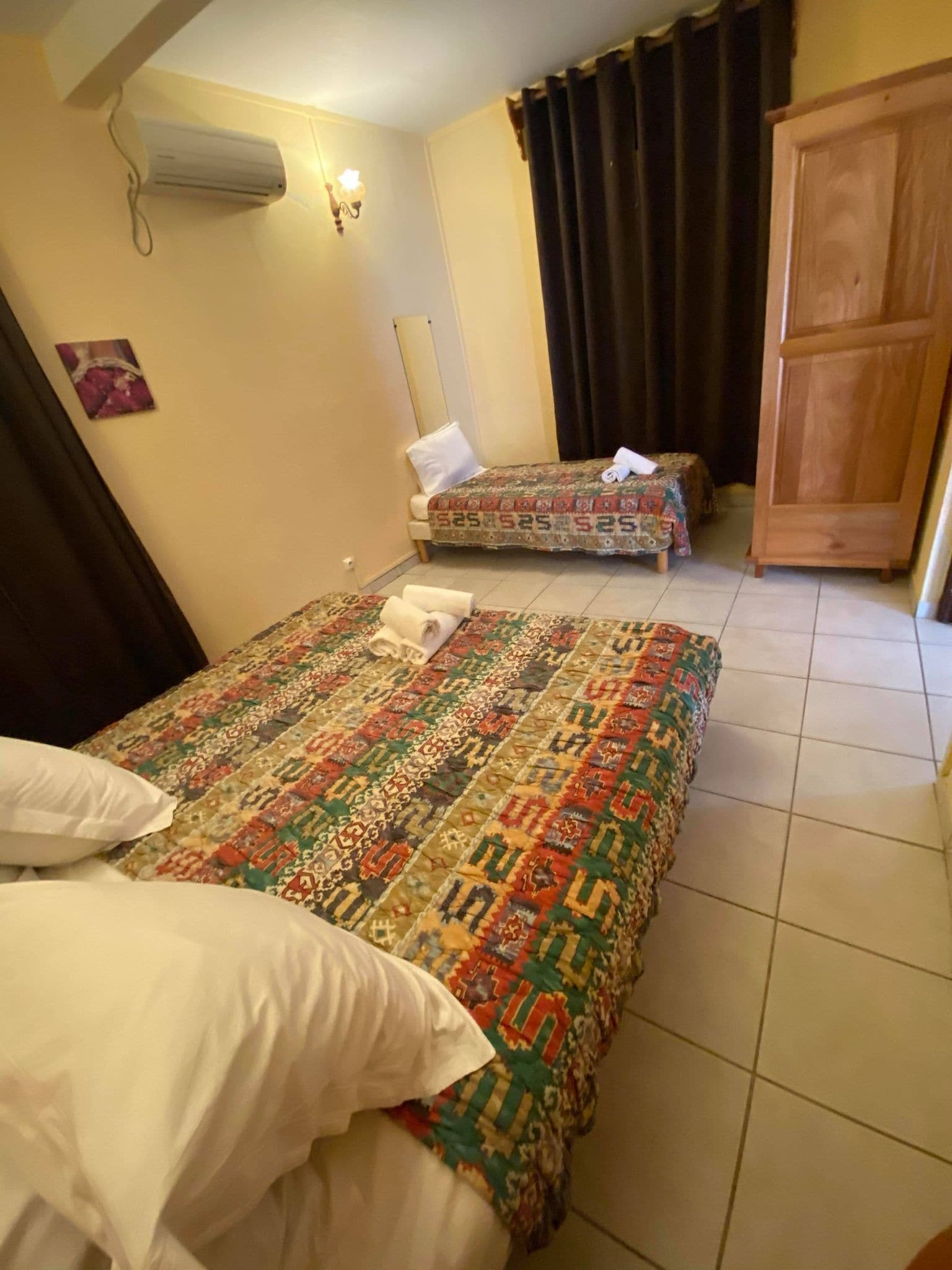 Habitación con dos camas, cada una con una colcha estampada y almohadas blancas. Se observa un armario de madera y un aire acondicionado de pared.
