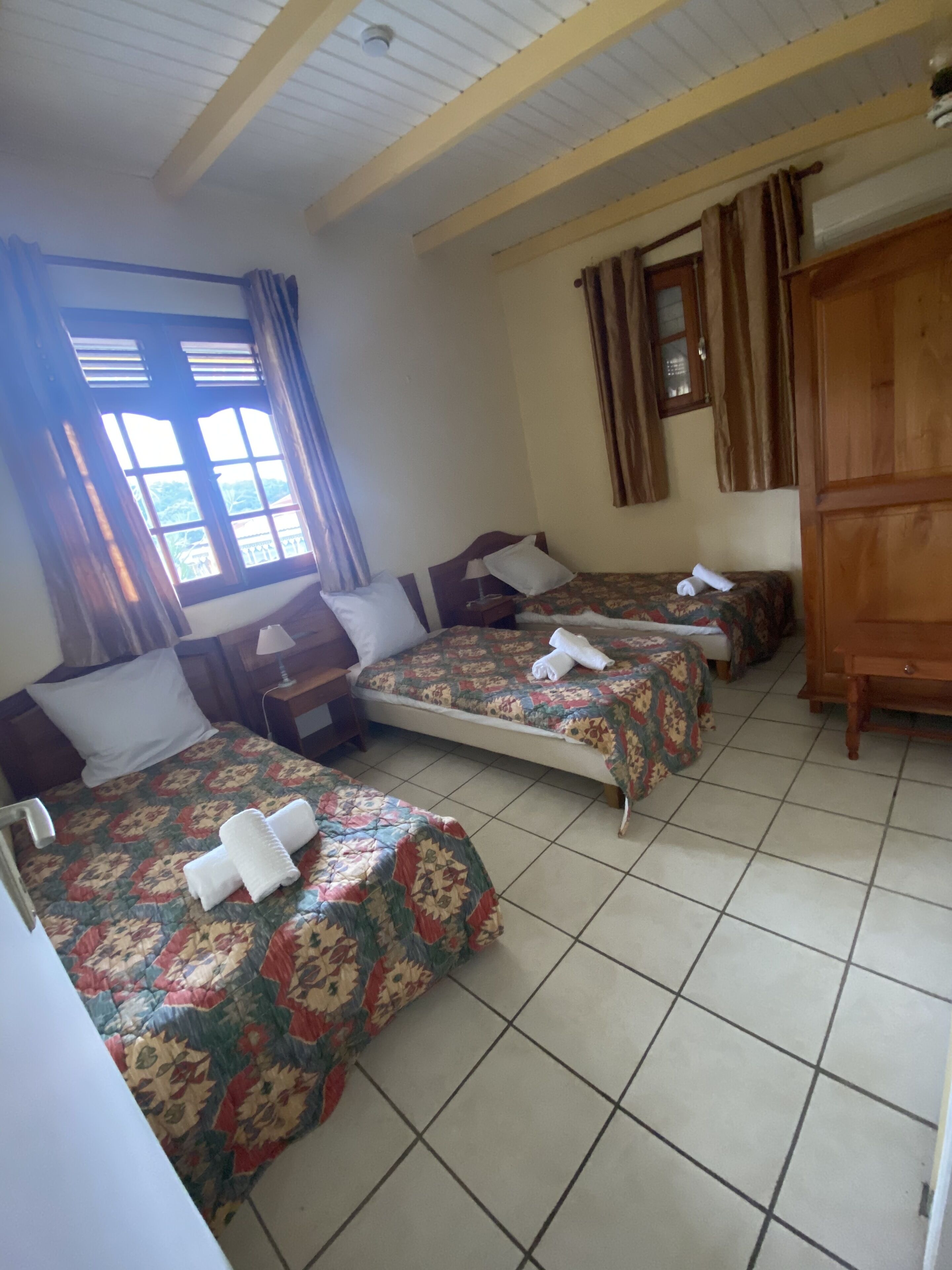Une chambre avec trois lits simples, chacun recouvert d’une couvre-lit à motifs floraux et agrémenté de serviettes blanches. On y trouve une fenêtre avec des rideaux, une armoire en bois et un sol carrelé.
