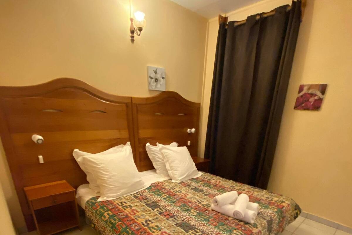 Una cama bien hecha con una colcha estampada, dos almohadas y un juego de toallas sobre la cama. Se observa un cabecero de madera, una mesita de noche con una lámpara y un reloj de pared. El cuarto tiene suelo de baldosas y una cortina oscura en la ventana.