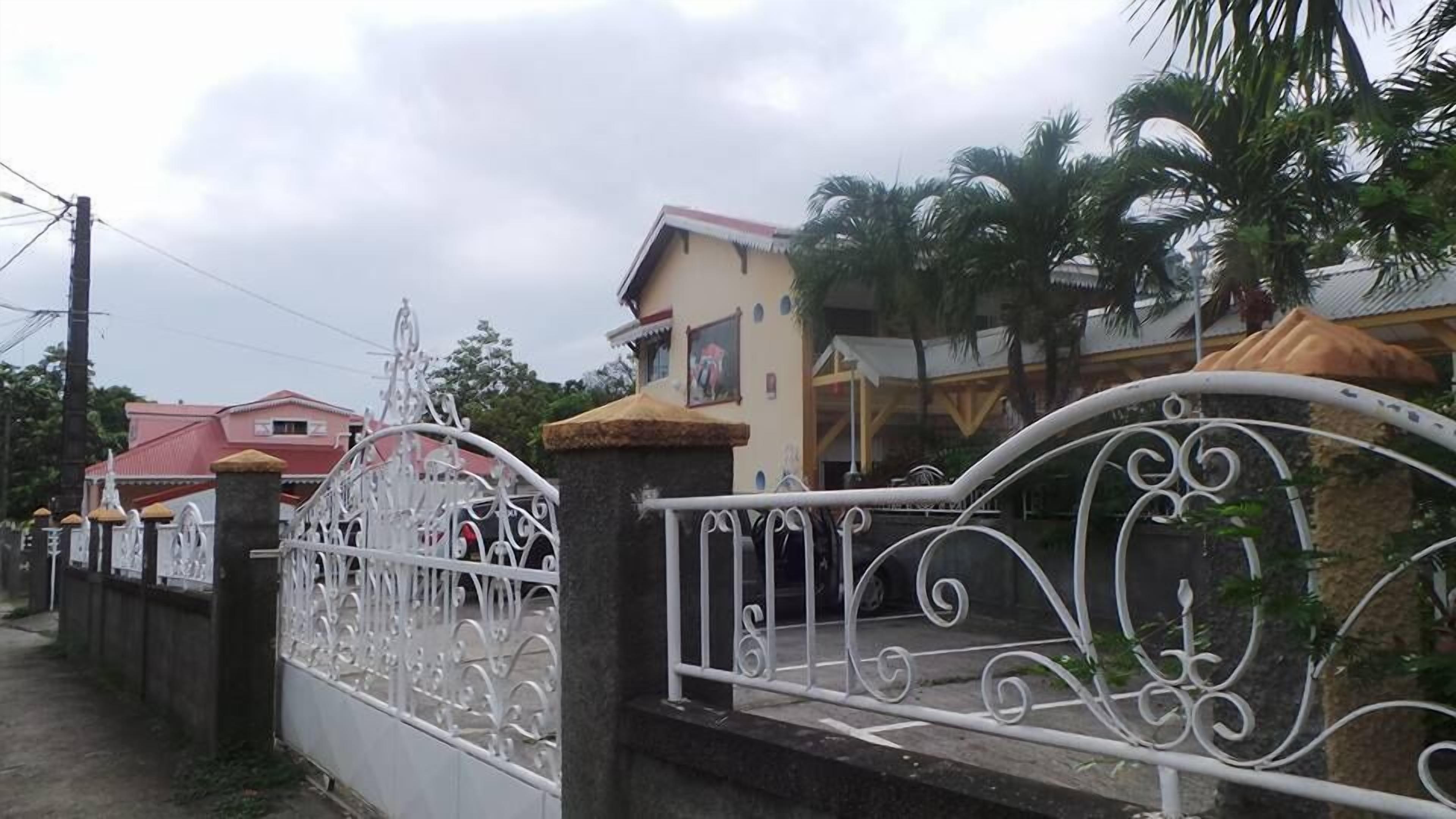 Área residencial murada com portão de ferro decorativo, caminho pavimentado e casas com telhados vermelhos.