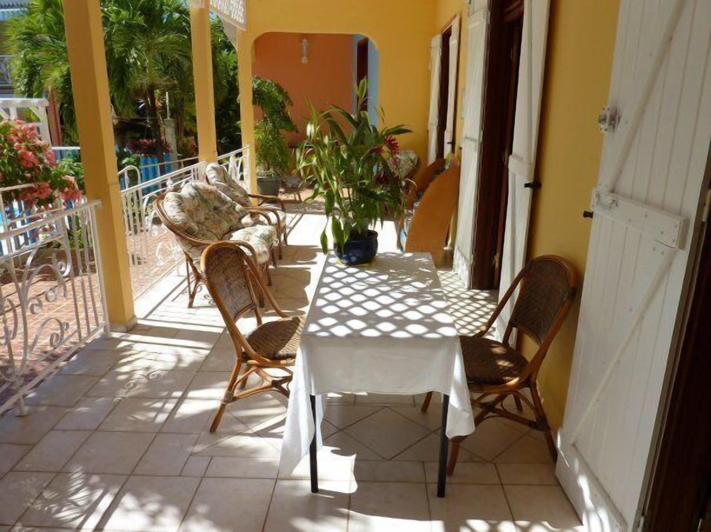 Een overdekt terras met een tafel en vier rieten stoelen, omgeven door potplanten en een gele muur.