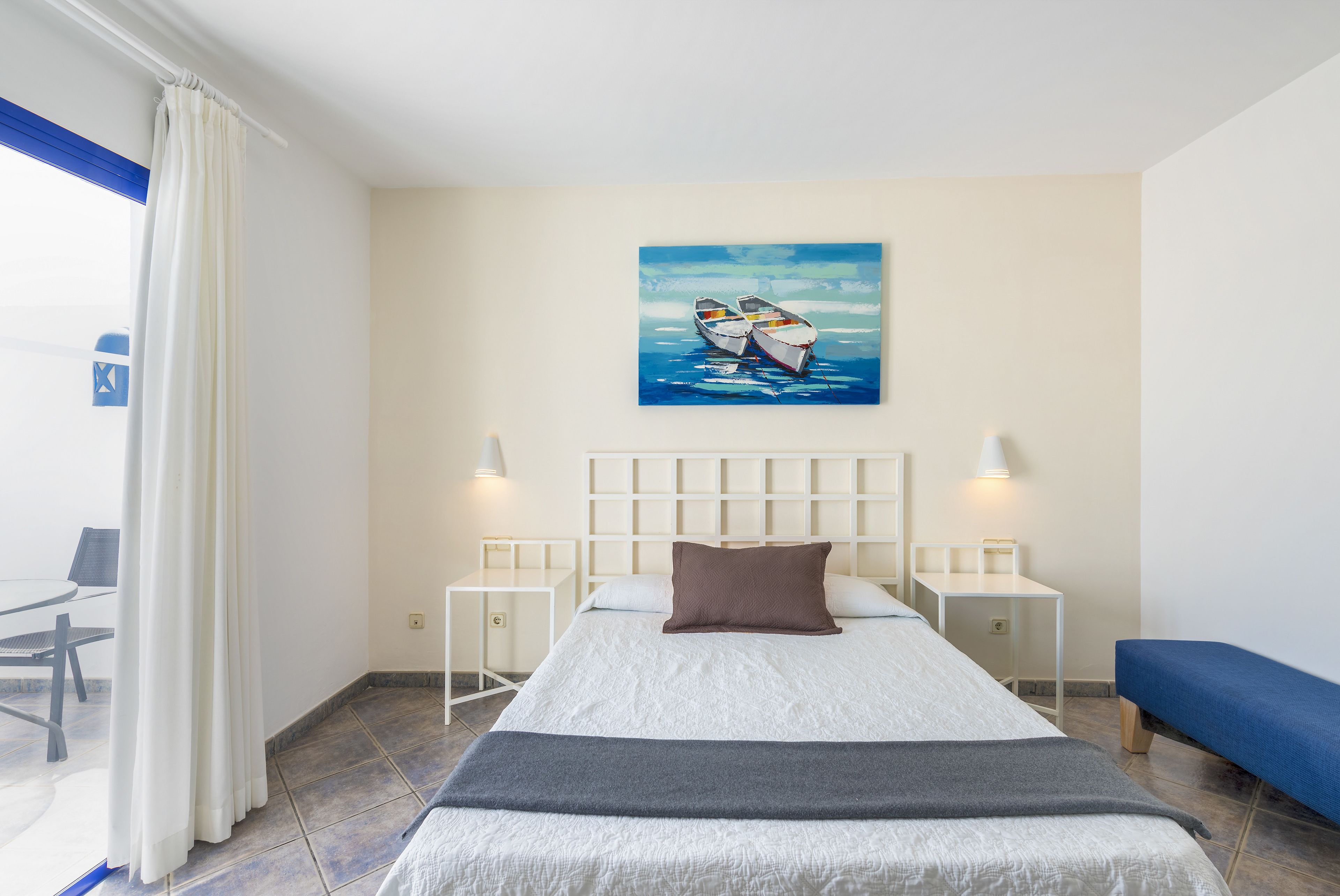 A hotel room with a bed, two bedside tables, a bench, a painting of boats on the wall, and a window with curtains.