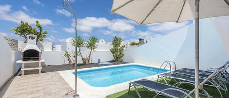Une piscine avec des chaises longues et un parasol, entourée d’une terrasse agrémentée d’un barbecue et de plantes.