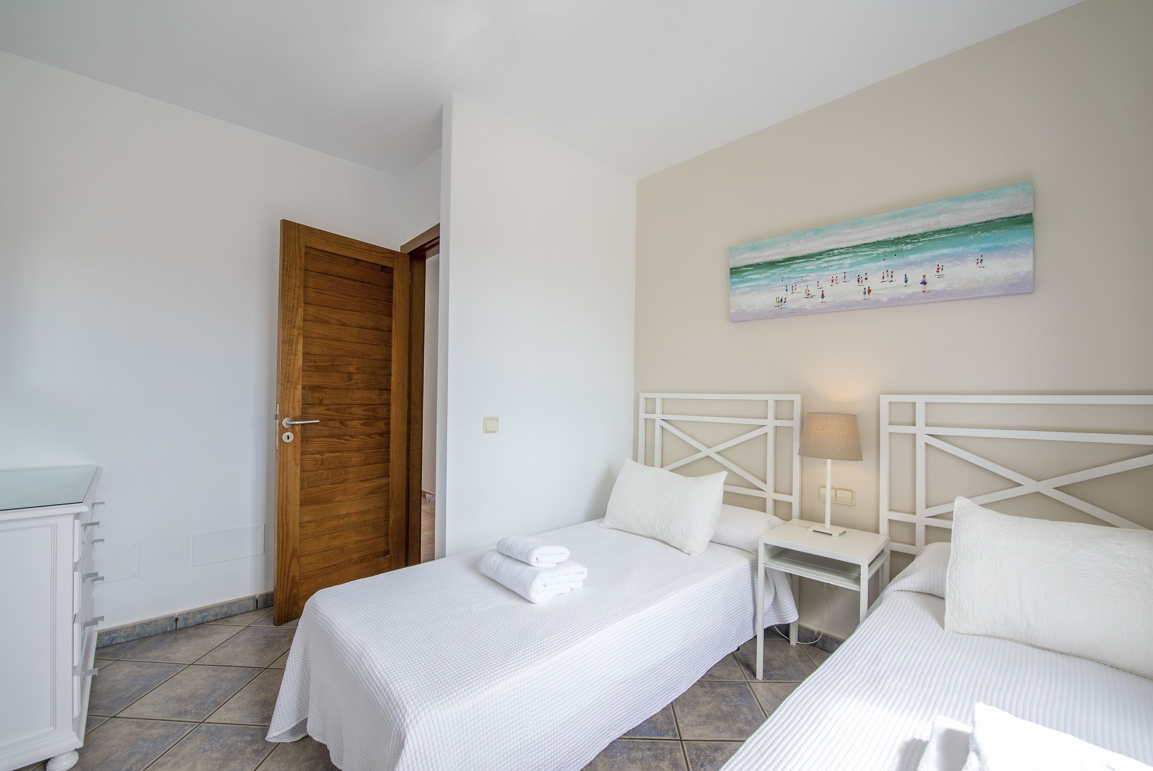 A hotel room with two beds, a wooden door, a painting of a beach scene, and a small bedside table with a lamp.