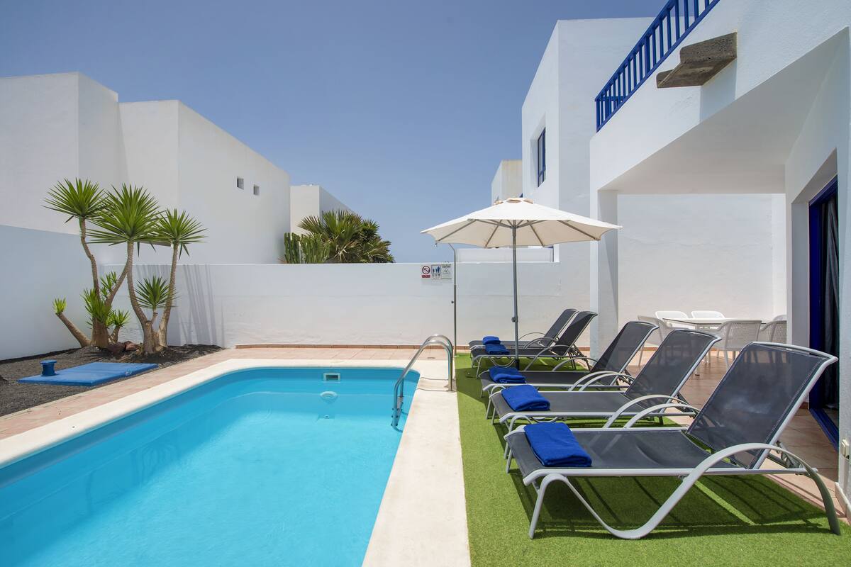 Piscina con sillas de descanso y una sombrilla, rodeada de paredes blancas y una barandilla azul.