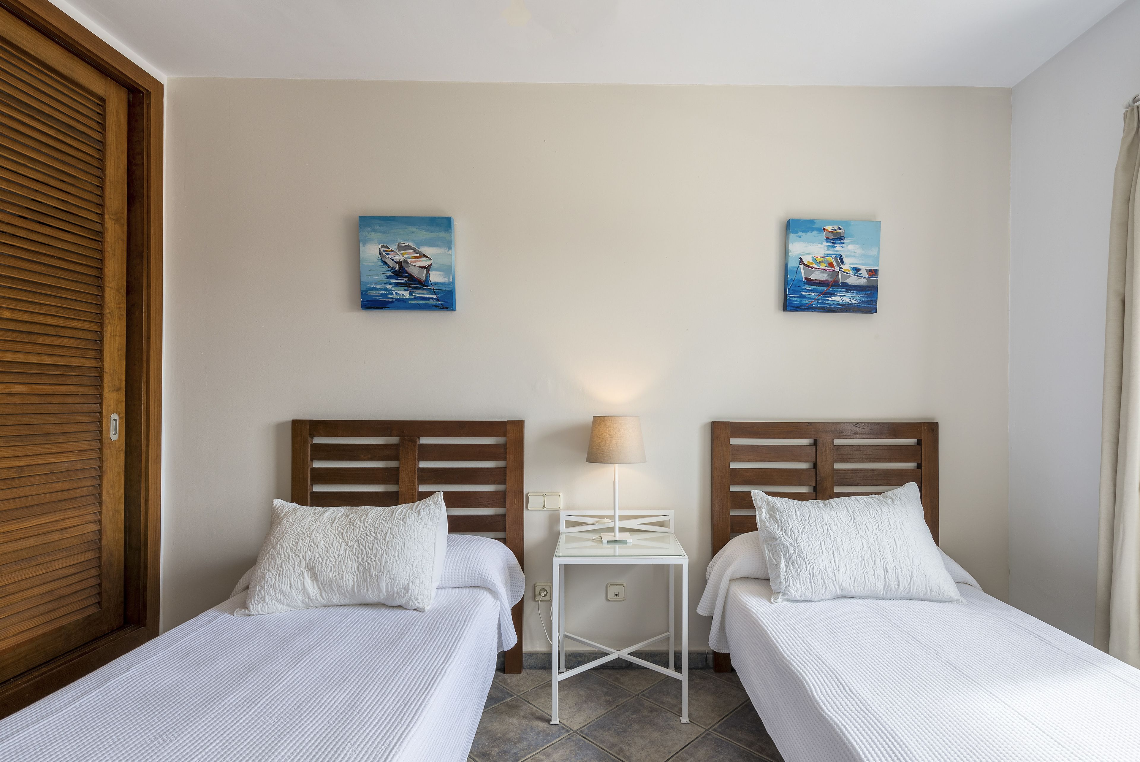 Une chambre d’hôtel avec deux lits simples, des têtes de lit en bois, une petite table de chevet blanche avec une lampe, et deux tableaux accrochés au mur.