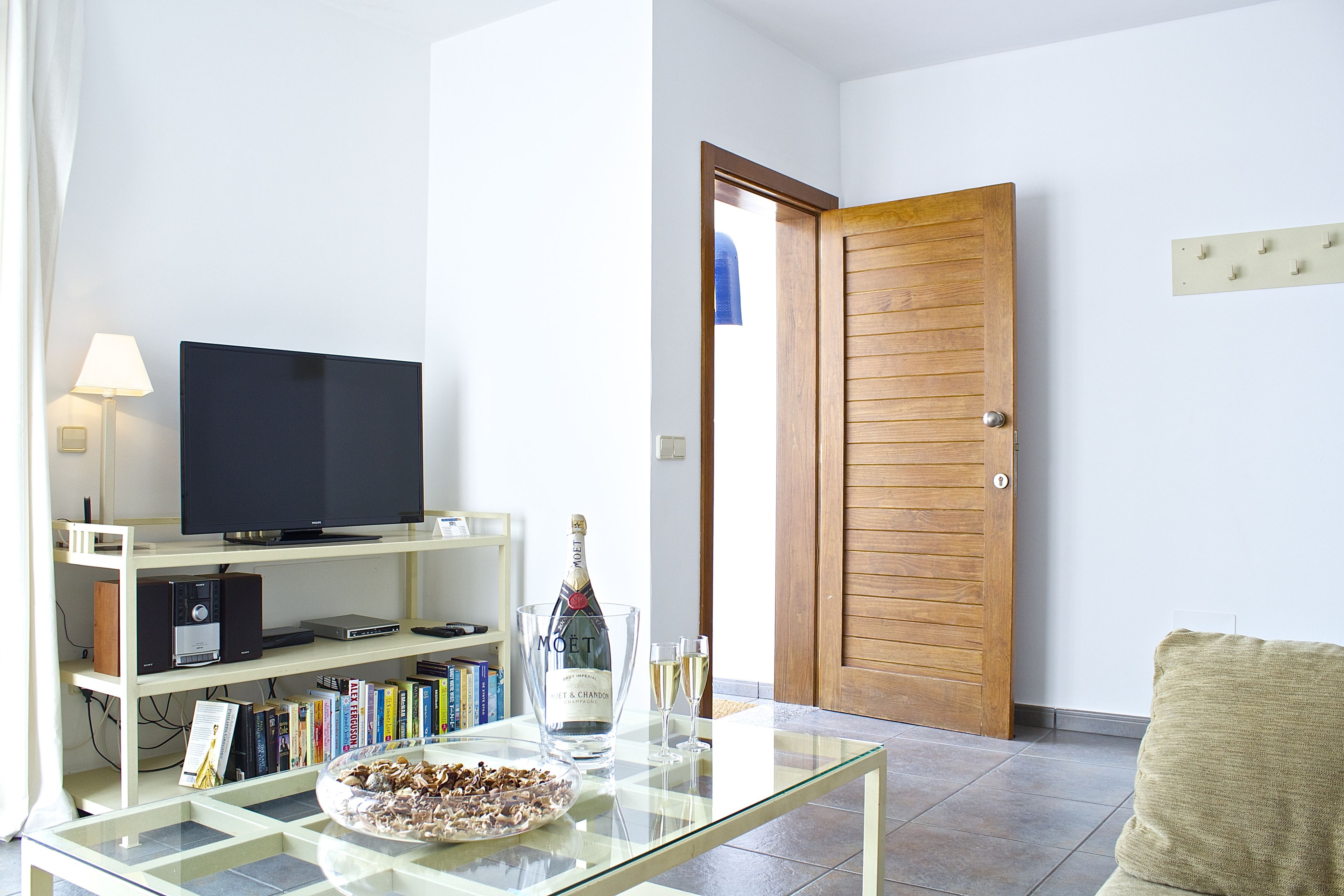 A living room with a television on a stand, a glass table with a bottle and glasses, and a bookshelf with books.