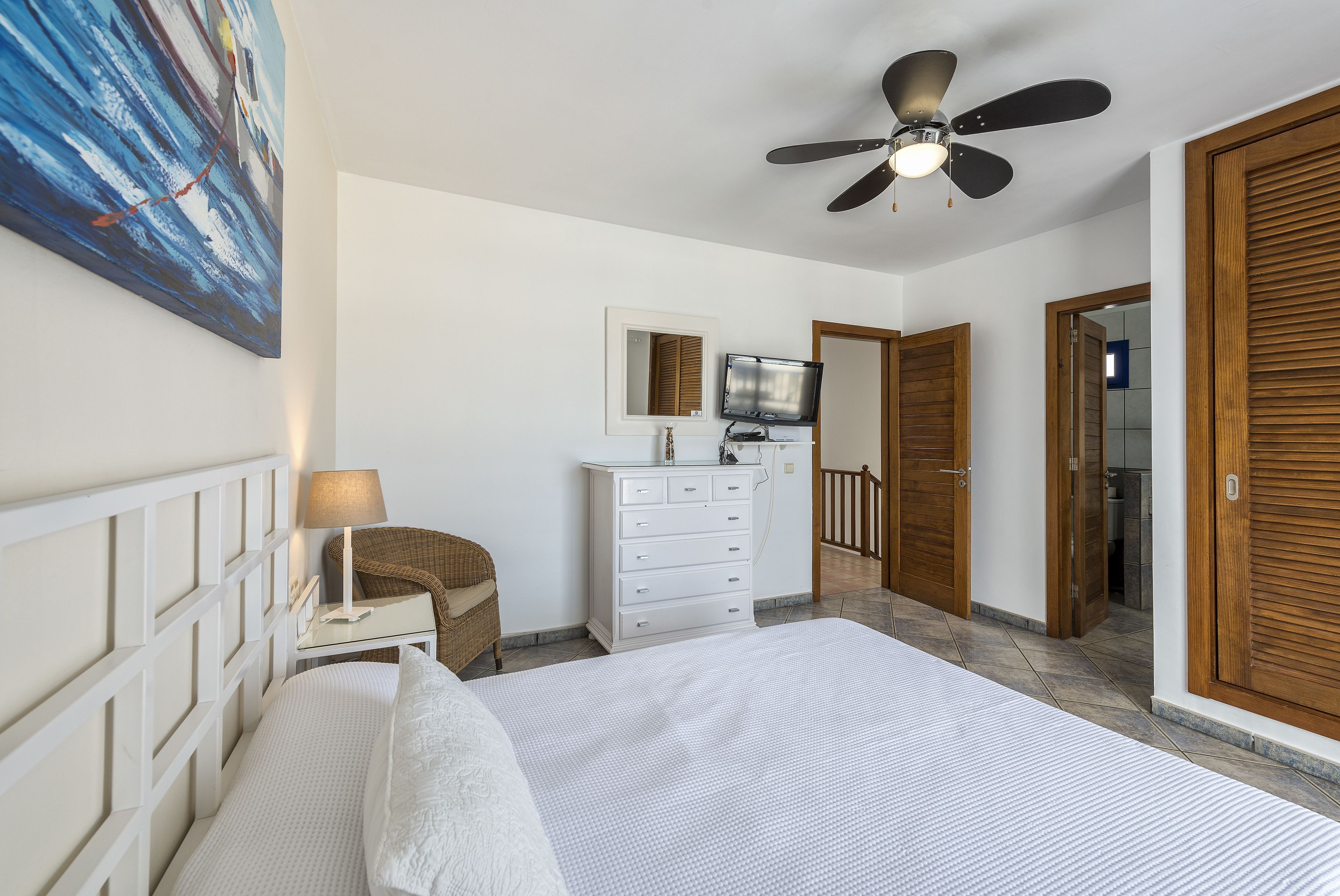 Une chambre à coucher comprenant un lit, un ventilateur de plafond, une télévision, une commode, une chaise et un tableau accroché au mur.