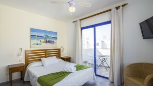 Habitación de hotel con cama, un cuadro de una playa, ventilador de techo, televisor y vista al exterior.