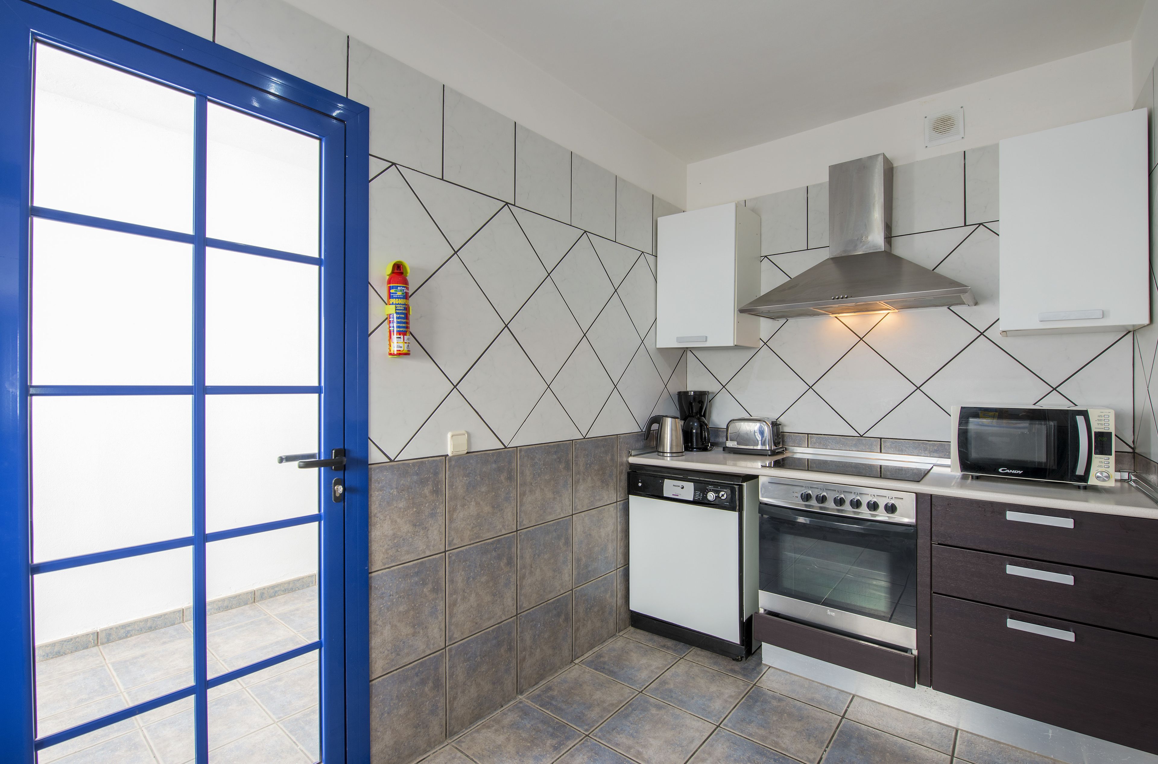 Una cocina con puerta azul, baldosas blancas y grises, estufa, microondas y lavavajillas.