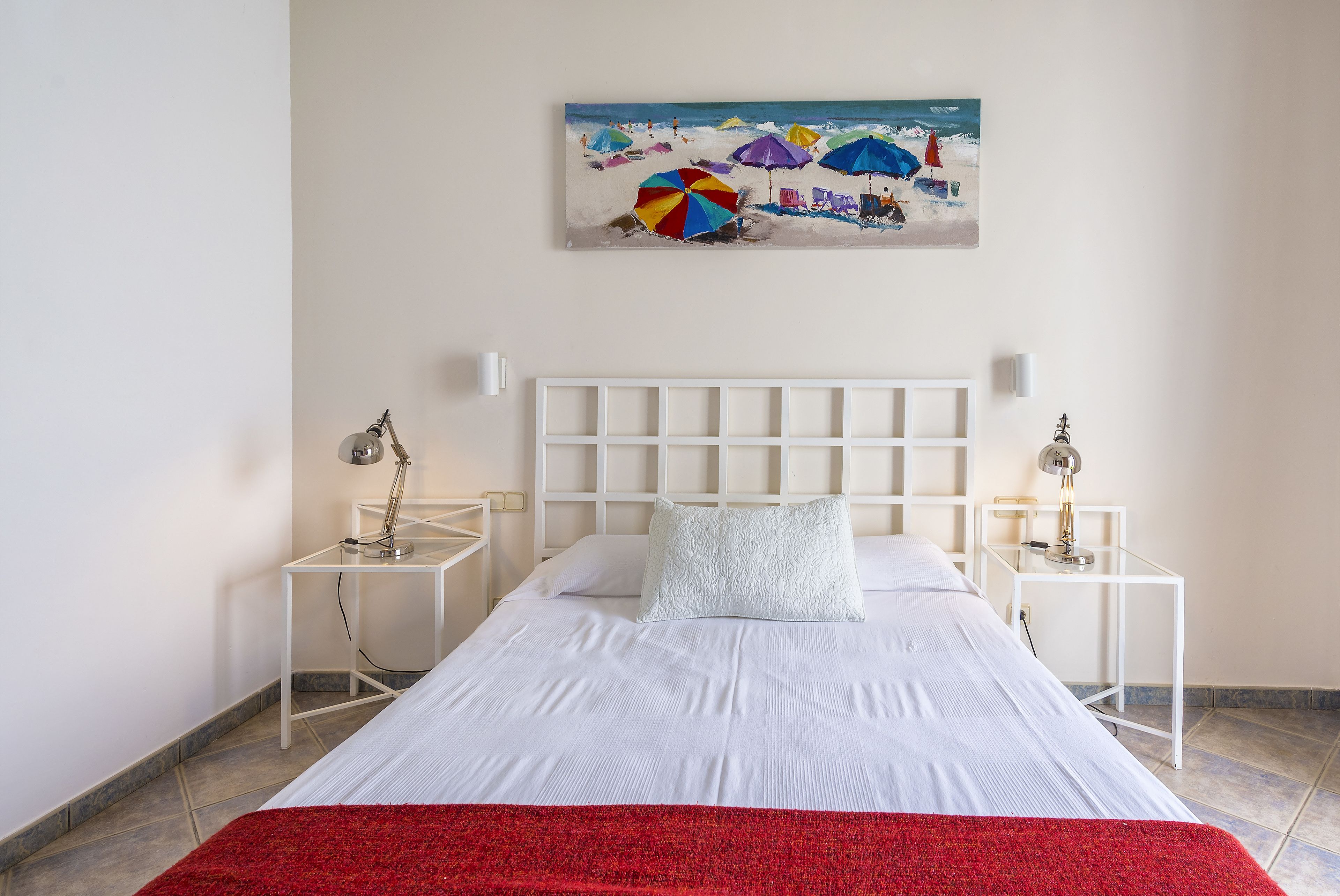 Habitación de hotel ordenada con cama, cabecera blanca, dos mesitas de noche con lámparas y un cuadro con motivos playeros en la pared.