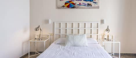 Habitación de hotel ordenada con cama, cabecera blanca, dos mesitas de noche con lámparas y un cuadro con motivos playeros en la pared.