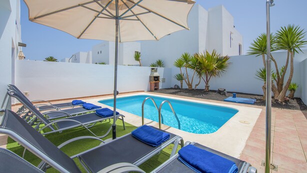 Un área de piscina con sillones de descanso, una sombrilla grande y una piscina azul cristalina.