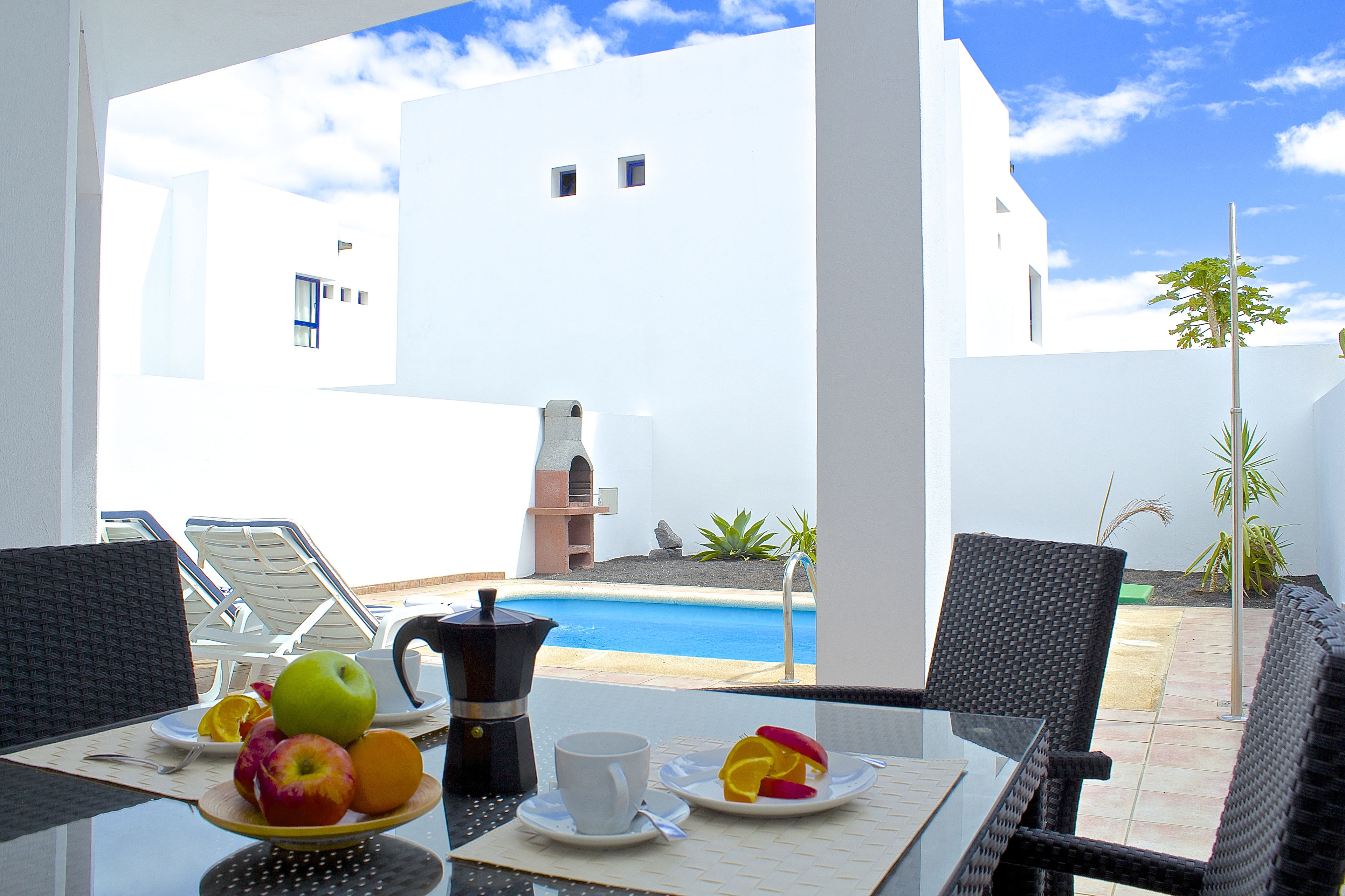 A table with breakfast items, a view of a swimming pool, and a white building in the background.