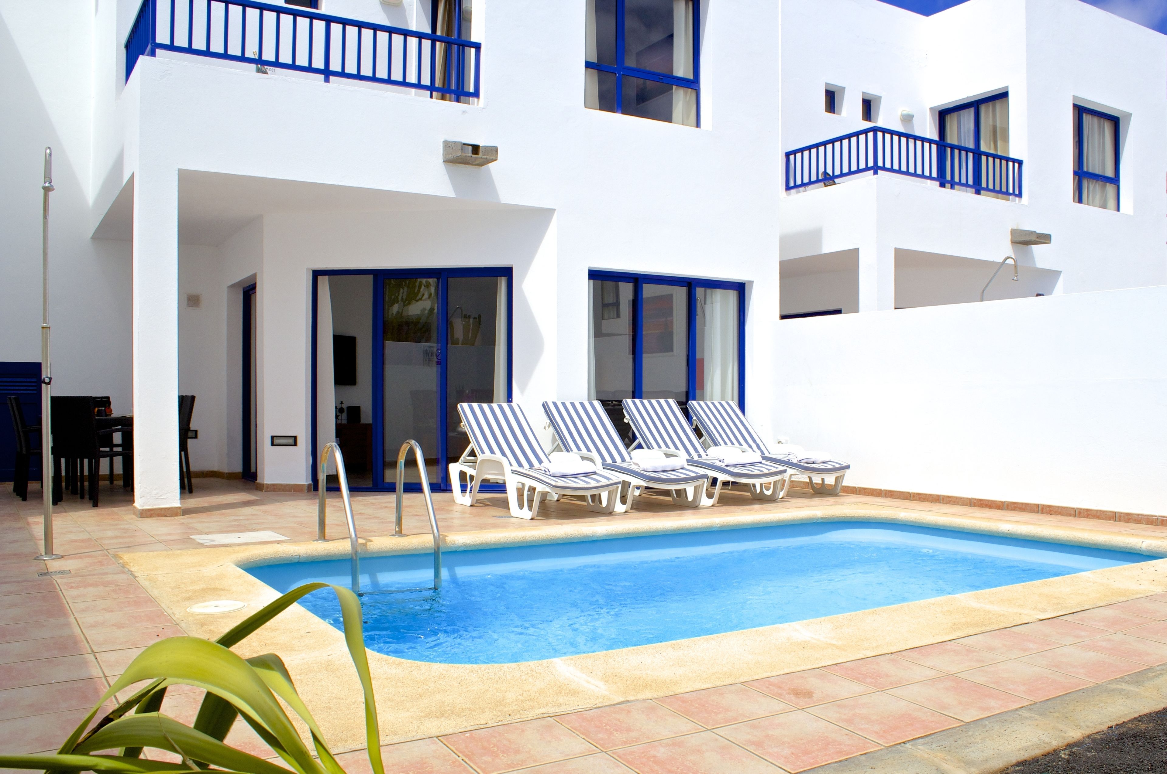 Zona de piscina con una piscina azul cristalina, paredes blancas y ventanas con marcos azules. Hay sillas de descanso y una puerta que da a un área cubierta.
