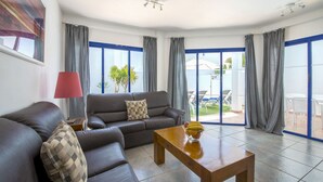 Una sala de estar moderna con un sofá, una mesita de centro y una lámpara. Cuenta con ventanas grandes con cortinas, un ventilador de techo y vistas al exterior.
