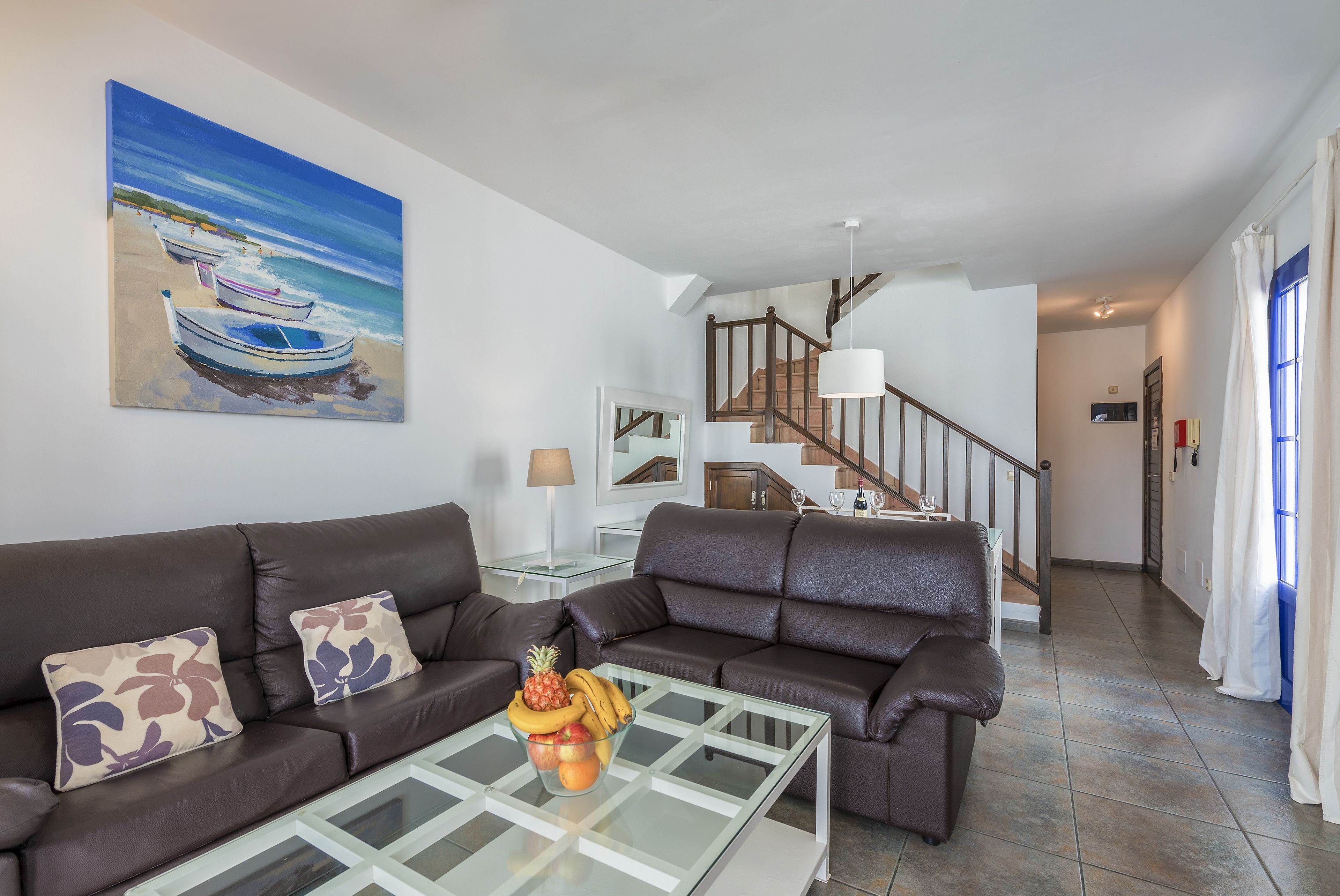 A living room with a glass coffee table, two brown leather sofas, a painting of boats on a beach, and a staircase leading to another room.