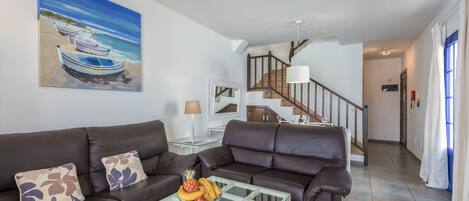 A living room with a glass coffee table, two brown leather sofas, a painting of boats on a beach, and a staircase leading to another room.