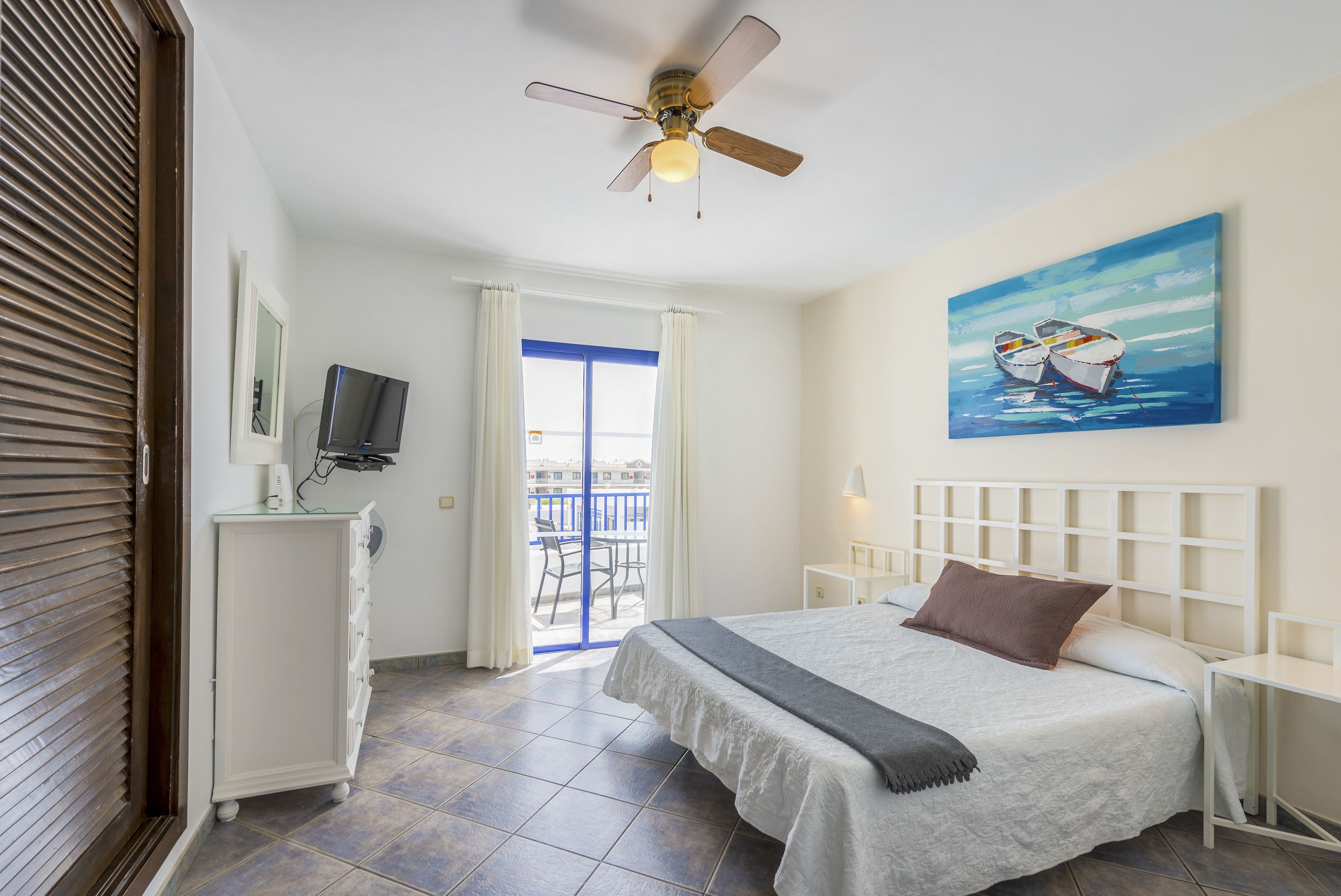 A bedroom with a bed, a ceiling fan, a painting of boats, a TV, and a balcony with a view.