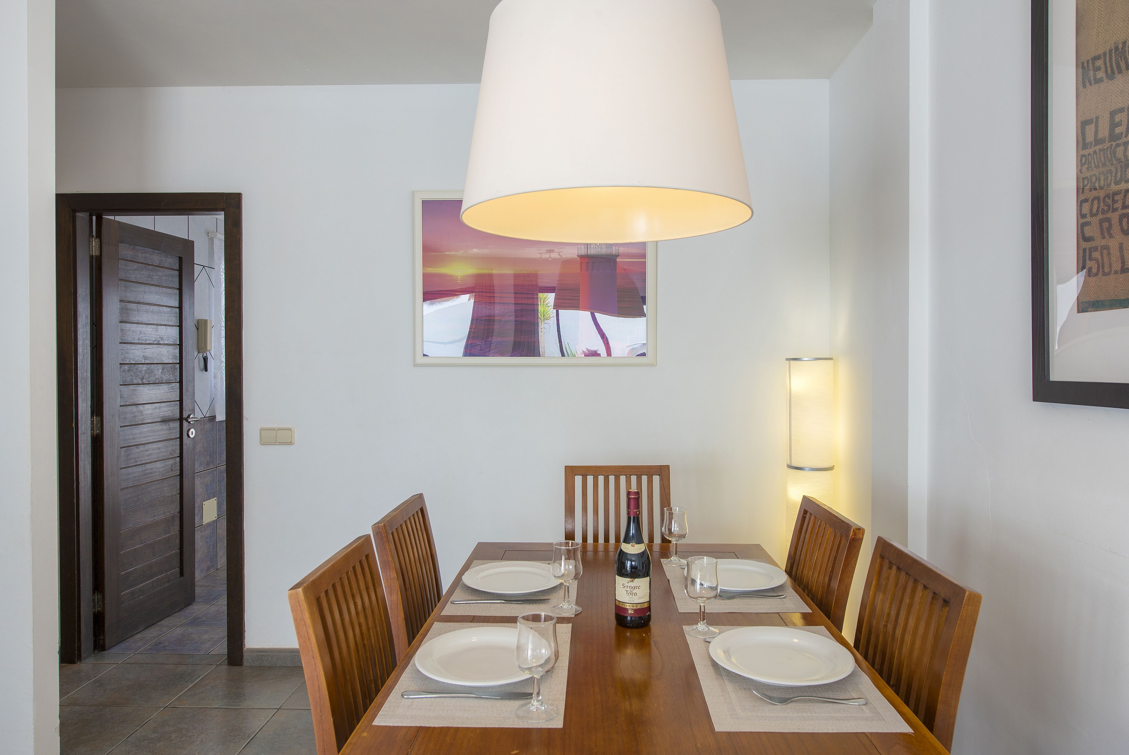 Un comedor con una mesa de madera dispuesta para cuatro, una botella de vino y un cuadro en la pared.