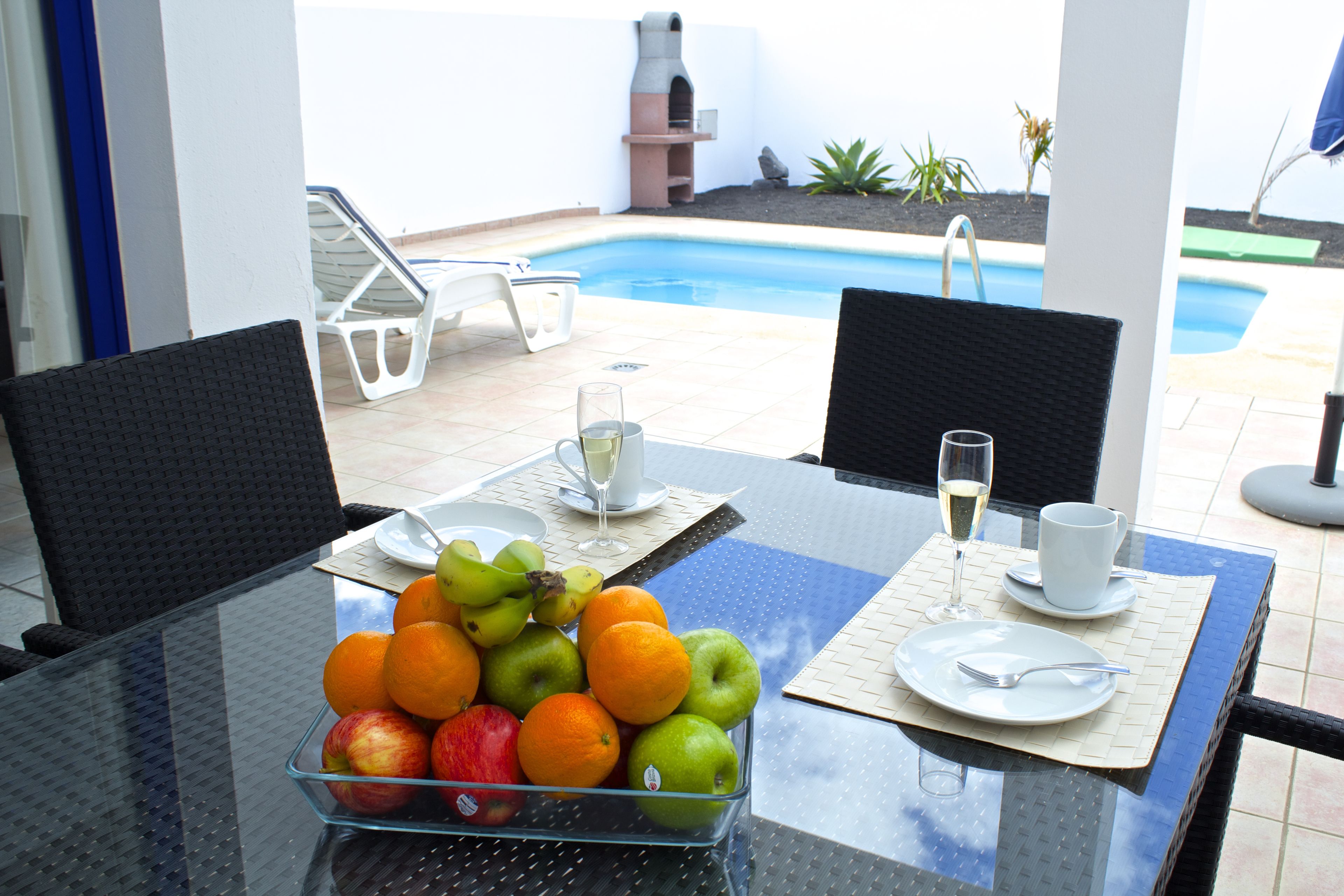 Una mesa de vidrio con un frutero, una copa de vino blanco y una taza sobre un mantel azul.