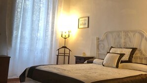 Ein Schlafzimmer mit Holzbalkendecke, einem Bett mit weißem Kopfteil, einem Nachttisch mit Lampe und einem Fenster mit Vorhängen.
