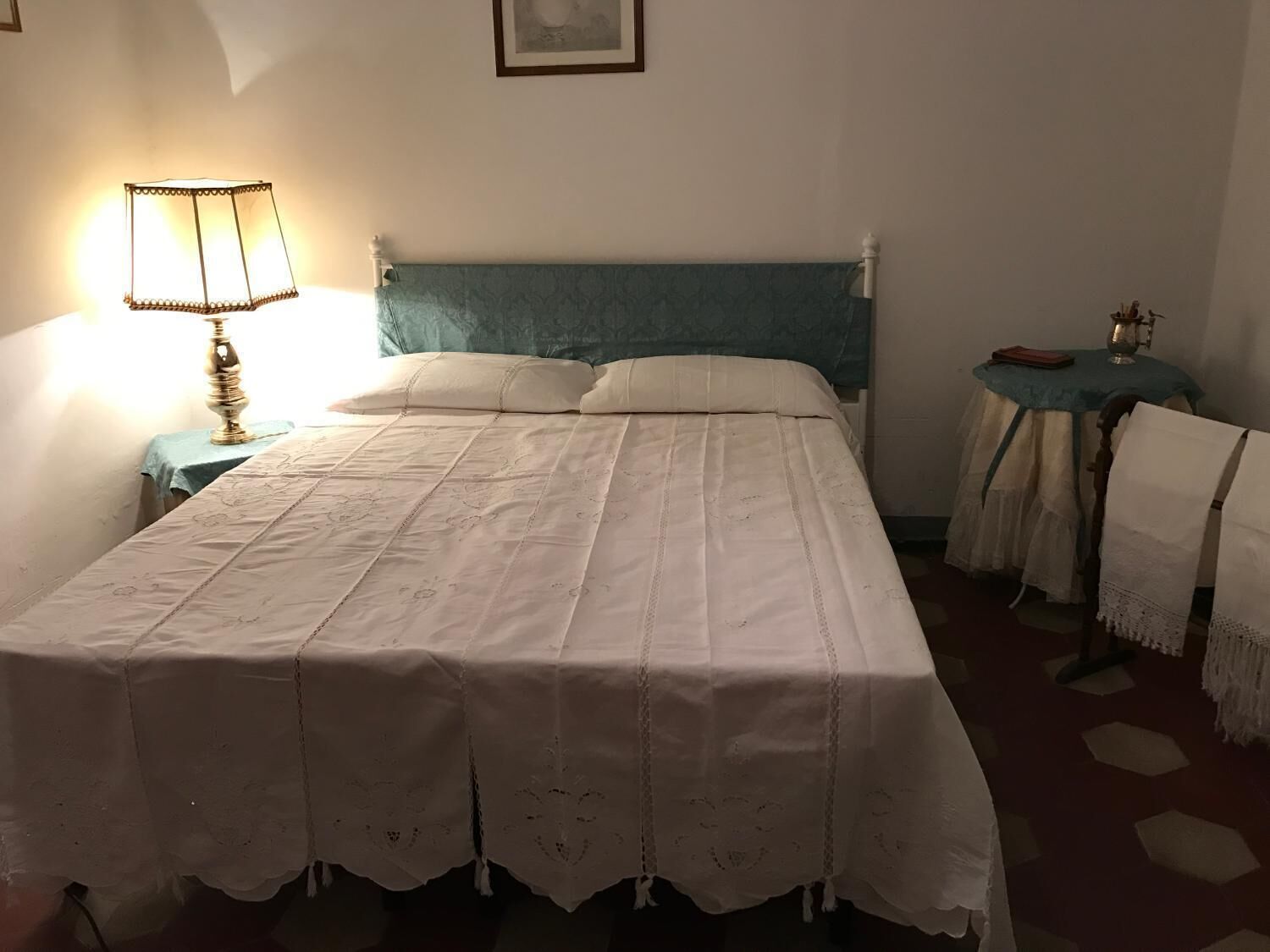 A neatly made bed with a white bedspread and matching pillows, a bedside lamp, and a small table with a book and a glass.