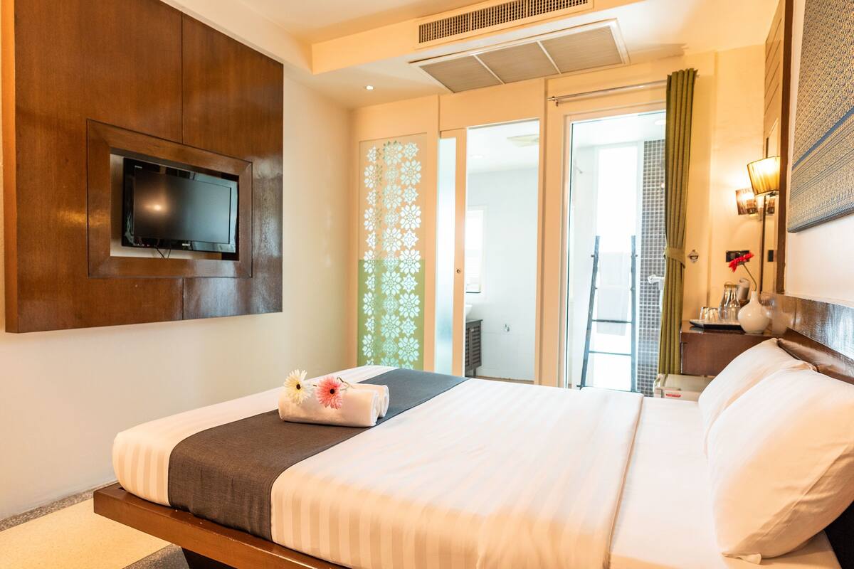 A hotel room with a bed, a TV mounted on the wall, a nightstand with a lamp, and a view of the outside through a glass door.