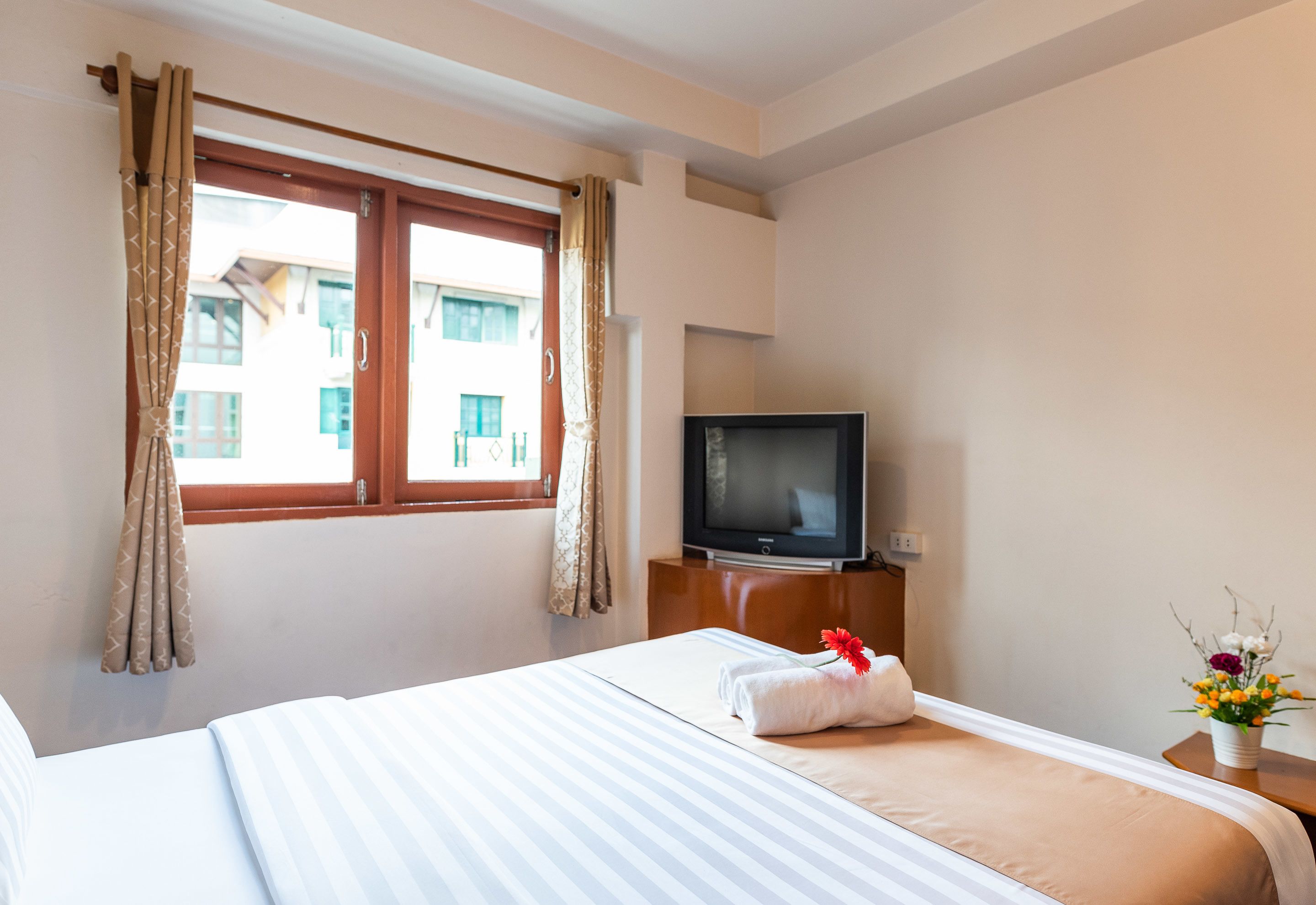 Una habitación de hotel con una cama, televisión, ventana con cortinas y un florero con flores.
