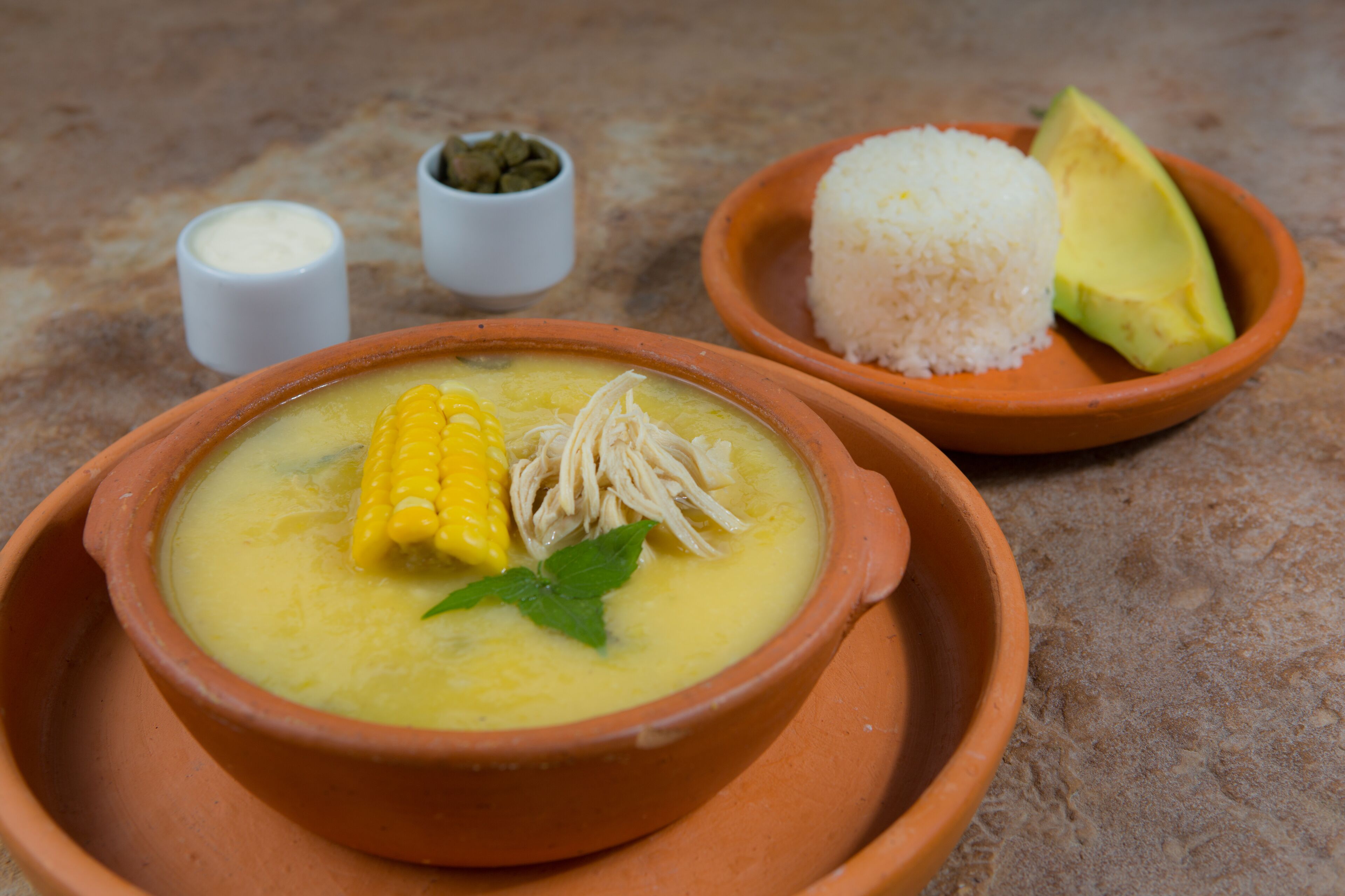 Two clay bowls with food, one containing a yellow soup with corn and mint, and the other with white rice and avocado.