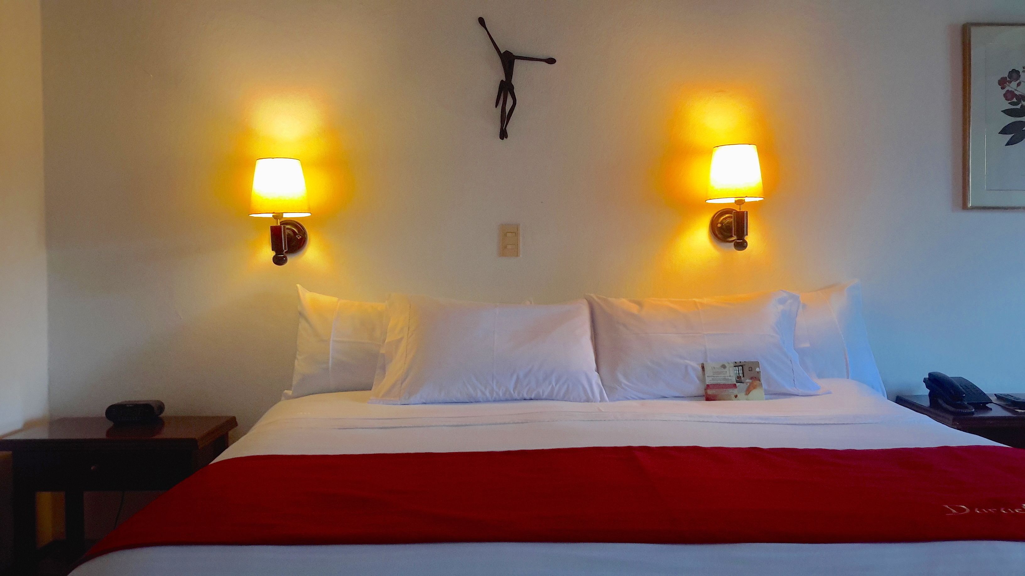 A hotel room with a bed, two wall-mounted lamps, a clock, and a framed picture on the wall.