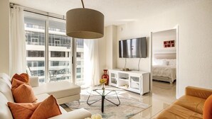 A modern living room with a sofa, armchair, and coffee table. A television is mounted on the wall, and a bed is visible in the background.