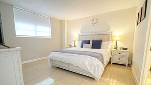 A bedroom with a large bed, two bedside tables with lamps, a window with blinds, and a decorative wall piece above the bed.